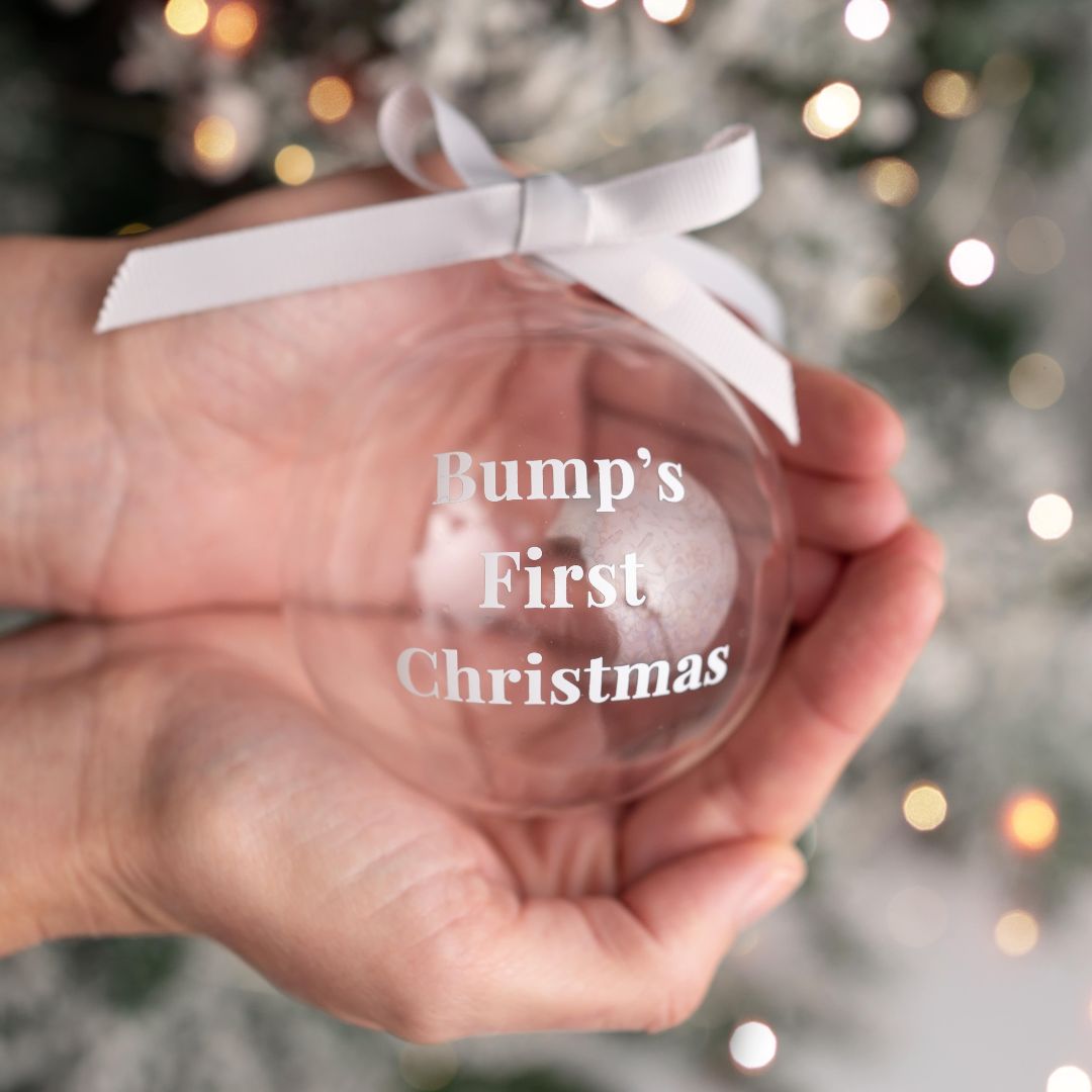 Glass bauble with white ribbon and “Bump’s First Christmas” text, held in two hands with a soft festive background.