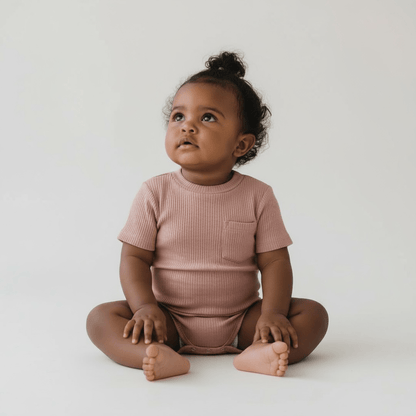 A baby wearing a warm taupe ribbed baby romper with short sleeves, pocket detail, and popper fastening.