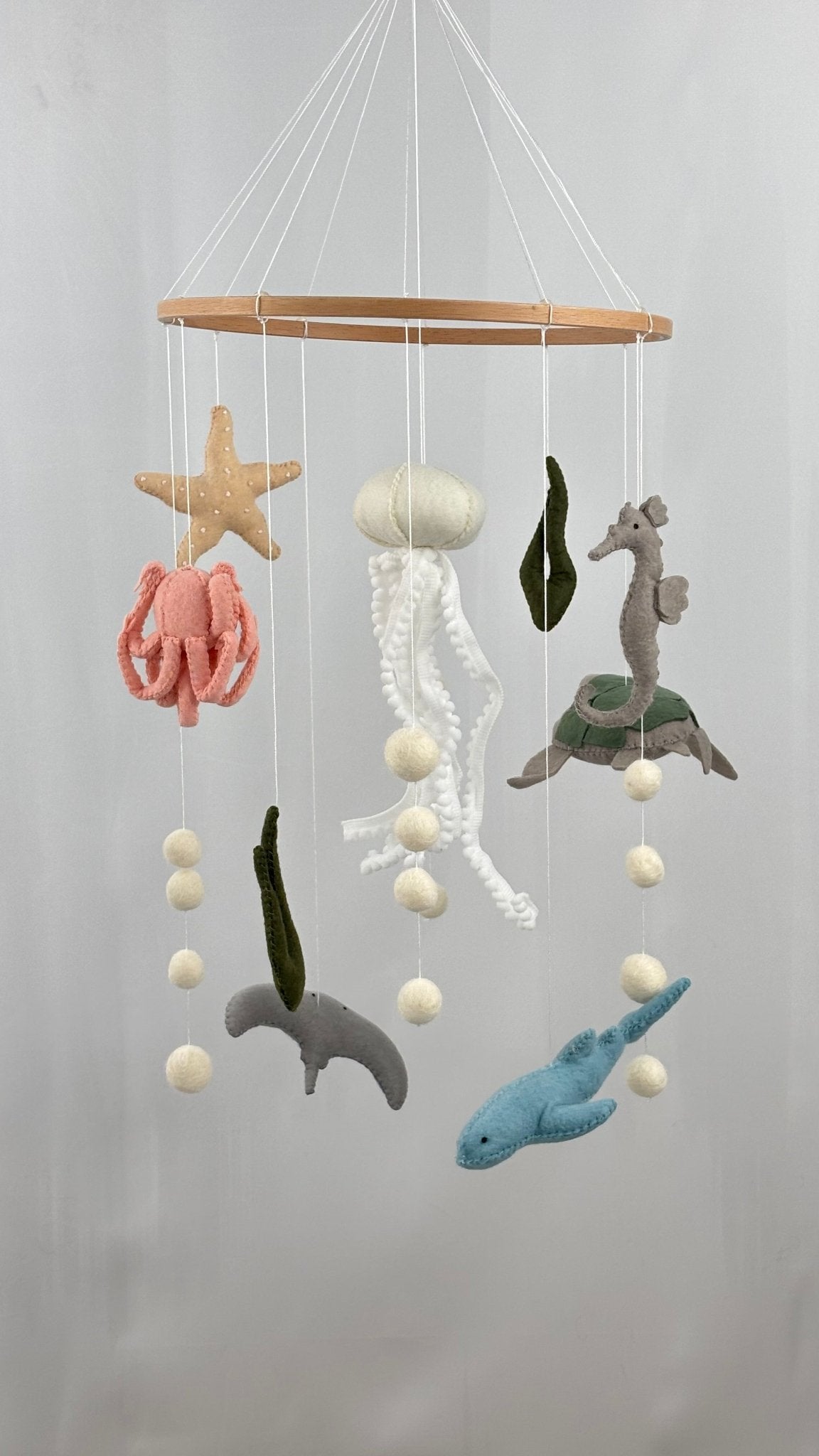 Hand-felted under-the-sea cot mobile with jellyfish, seahorse, octopus, starfish, turtle, stingray, and shark hanging from a wooden ring, 65 cm drop, on white background.